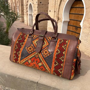 May include: A brown leather duffel bag with a colorful woven fabric panel. The fabric features geometric patterns in red, orange, and yellow. The bag has a top handle and a shoulder strap.