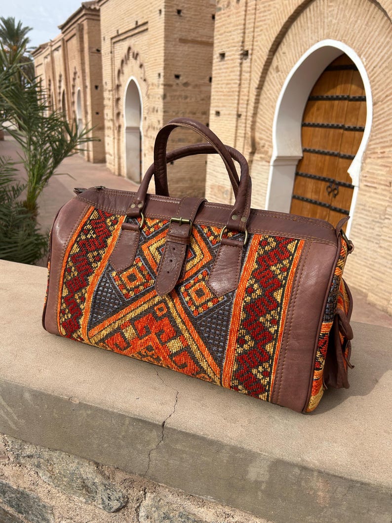 May include: A brown leather duffel bag with a colorful woven fabric panel. The fabric features geometric patterns in red, orange, and yellow. The bag has a top handle and a shoulder strap.