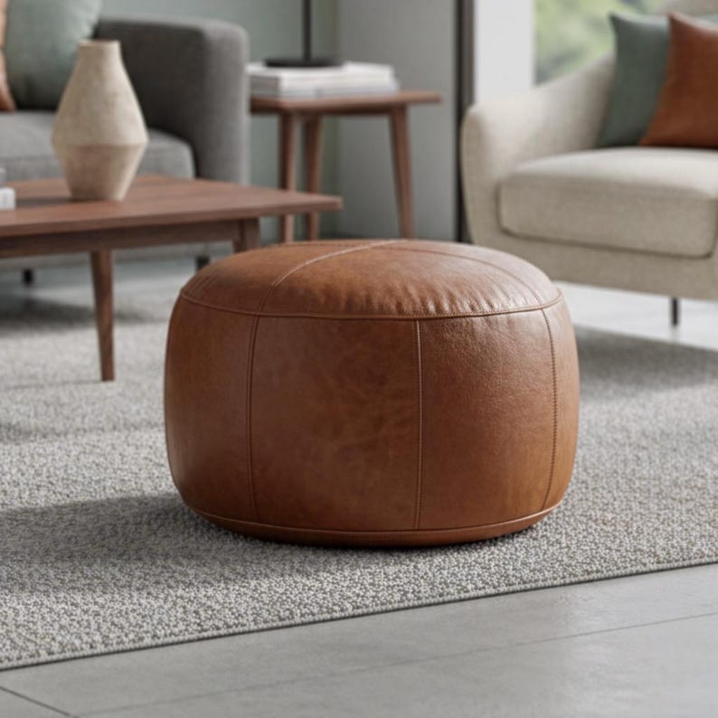 Peut inclure: Un pouf rond en cuir marron avec des coutures apparentes. Le pouf est pos&eacute; sur un tapis gris dans un salon. Ce meuble est un &eacute;l&eacute;ment neutre et chaleureux.