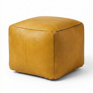 Peut inclure: Un pouf carré en cuir jaune moutarde. Le meuble en forme de cube présente des coutures et des bords arrondis. Le pouf est de couleur unie et repose sur un fond blanc. Ce meuble convient à un salon ou une chambre.