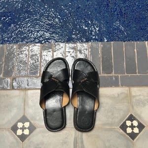 May include: A pair of black leather sandals with a cross-strap design. The sandals are placed on a gray tiled surface with a blue pool in the background. The sandals have a leather insole and a stitched design.