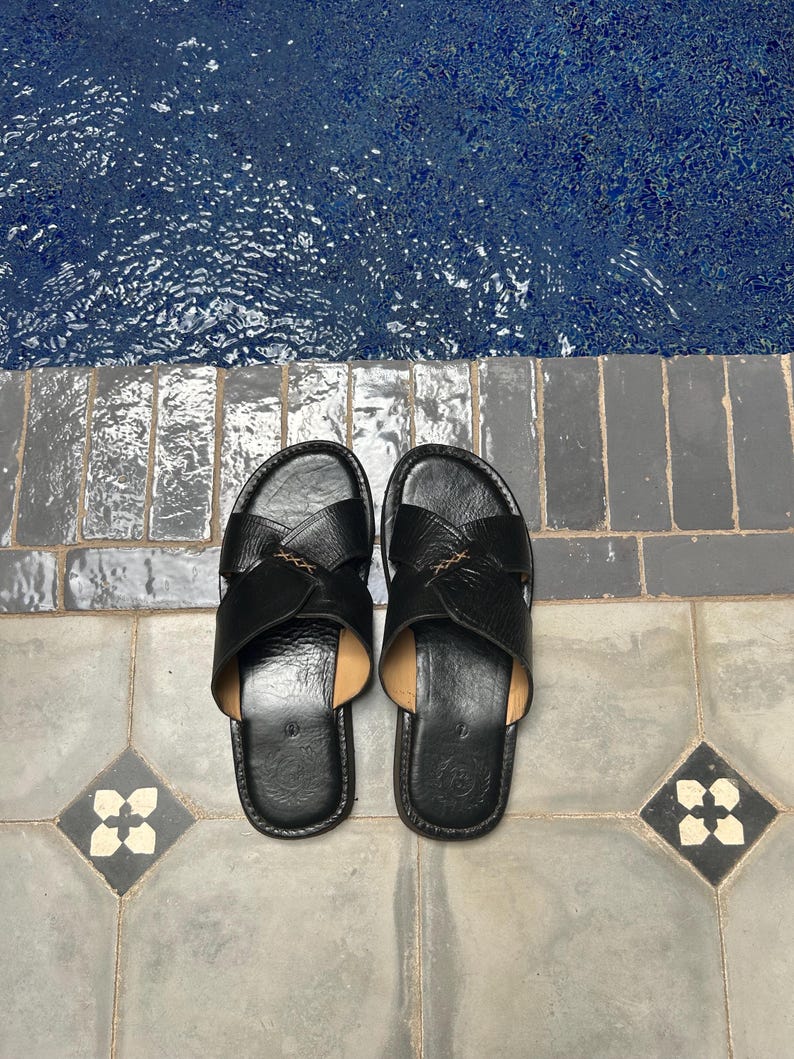 May include: A pair of black leather sandals with a cross-strap design. The sandals are placed on a gray tiled surface with a blue pool in the background. The sandals have a leather insole and a stitched design.