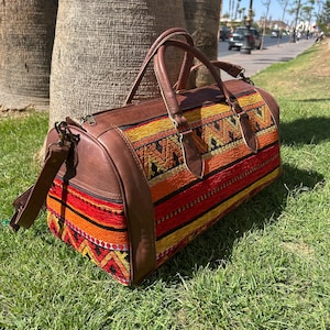 Puede incluir: Una bolsa de lona de cuero marrón y tela tejida con un colorido patrón geométrico en rojo, naranja y amarillo. La bolsa tiene un asa superior y una correa para el hombro, apoyada sobre hierba verde.
