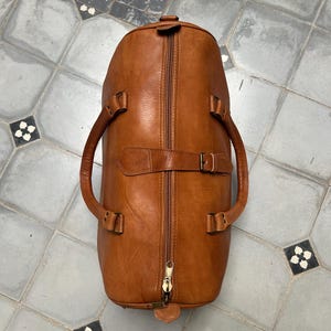 May include: A brown leather duffel bag with a full-length zipper and two handles. The bag has a buckle closure and a small leather tag at the base. The bag is made of a smooth, textured leather.