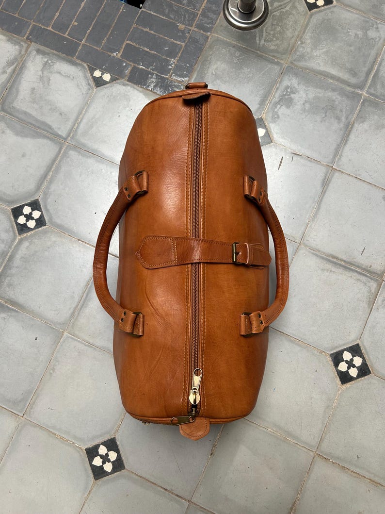 May include: A brown leather duffel bag with a full-length zipper and two handles. The bag has a buckle closure and a small leather tag at the base. The bag is made of a smooth, textured leather.