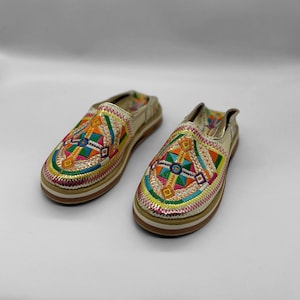 Puede incluir: Un par de zapatillas doradas bordadas con coloridos patrones geométricos. Las zapatillas tienen una suela de goma blanca.