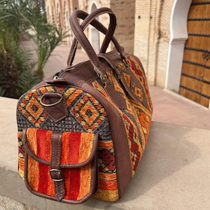 May include: A brown leather and woven fabric travel bag with a geometric pattern in orange, yellow, and red. The bag has a front pocket with a buckle closure and two top handles.