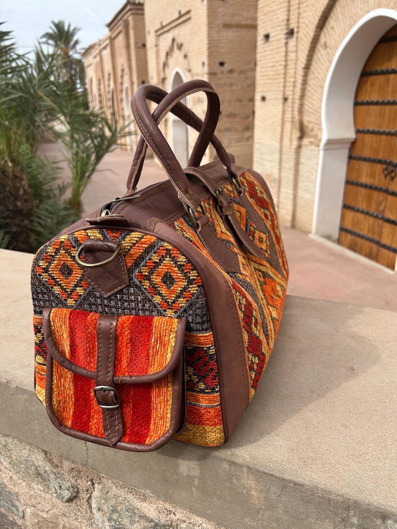 May include: A brown leather and woven fabric travel bag with a geometric pattern in orange, yellow, and red. The bag has a front pocket with a buckle closure and two top handles.