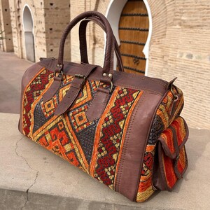 May include: A brown leather duffel bag with a colorful woven fabric panel. The fabric features geometric patterns in shades of orange, red, yellow, and black. The bag has a top handle and a side pocket.