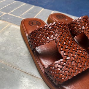May include: A pair of brown leather sandals with a woven strap design. The sandals have a flat sole and a logo embossed on the footbed. The sandals are made of leather and are designed for casual wear.