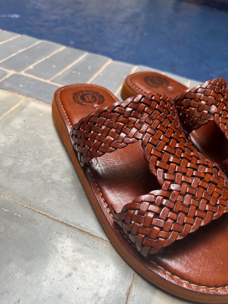 May include: A pair of brown leather sandals with a woven strap design. The sandals have a flat sole and a logo embossed on the footbed. The sandals are made of leather and are designed for casual wear.