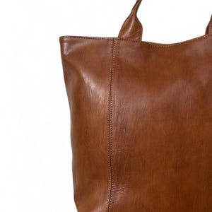 May include: A close-up of a brown leather tote bag with two handles. The bag has a rectangular shape with a vertical seam down the center. The leather has a smooth, slightly glossy finish.
