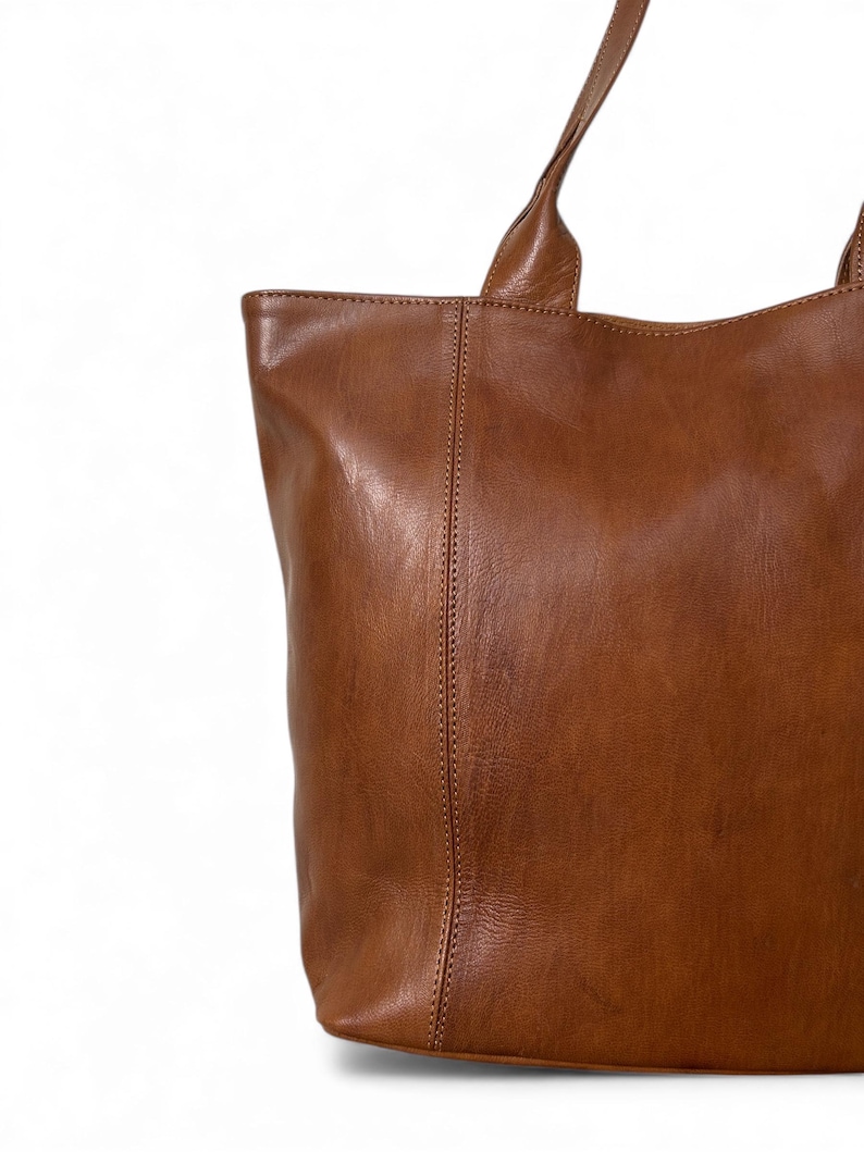 May include: A close-up of a brown leather tote bag with two handles. The bag has a rectangular shape with a vertical seam down the center. The leather has a smooth, slightly glossy finish.