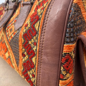 May include: A close-up of a duffel bag with a woven textile design in red, orange, and yellow. The bag features brown leather accents, including handles and trim. The bag has a rectangular shape.