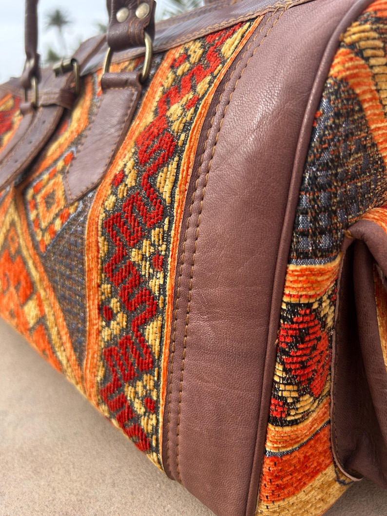 May include: A close-up of a duffel bag with a woven textile design in red, orange, and yellow. The bag features brown leather accents, including handles and trim. The bag has a rectangular shape.