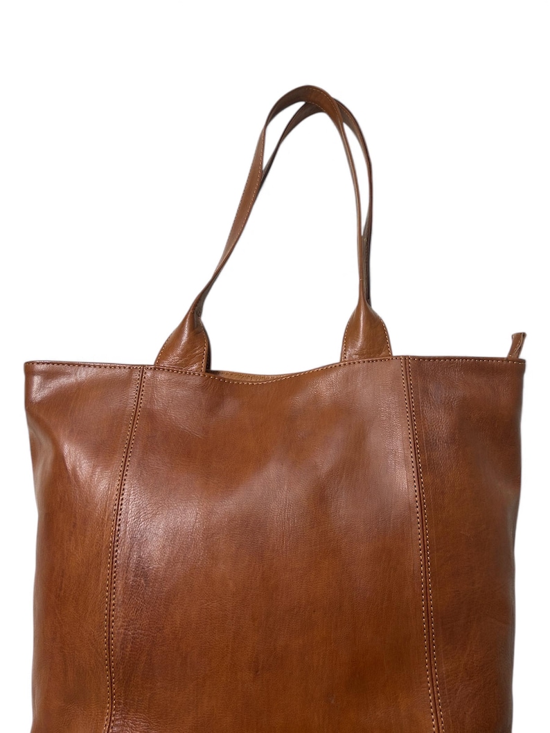 May include: A brown leather tote bag with two long handles. The bag has a rectangular shape with vertical seams on the front and back. The leather has a smooth texture and a warm, natural color.