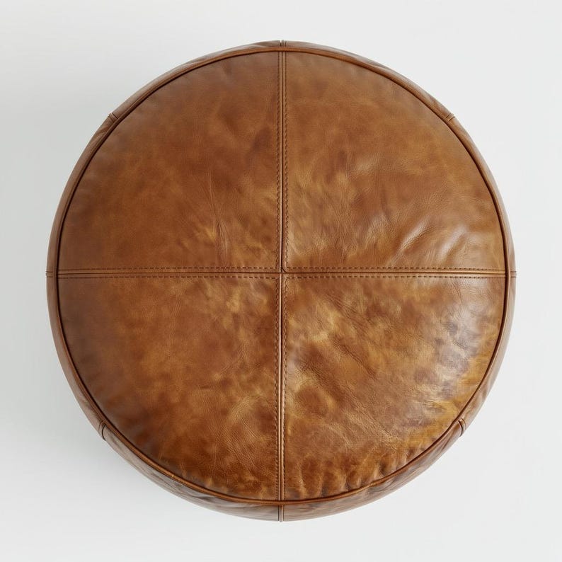 Peut inclure: Un pouf rond en cuir marron avec un design cousu. Le pouf est divis&eacute; en quatre sections par des coutures. Le cuir a un aspect l&eacute;g&egrave;rement vieilli, avec des variations de couleur et de texture.