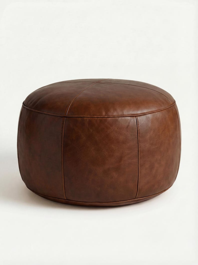 May include: A brown leather ottoman with a rounded, cylindrical shape. The ottoman has a stitched seam detail and is made of a smooth, dark brown leather. It is a comfortable and stylish piece of furniture.