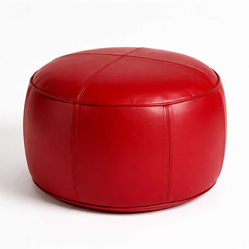 May include: A round, red leather ottoman with a stitched cross pattern on top. The ottoman is a solid, vibrant red color and has a cylindrical shape. The seams are visible, adding detail to the design. It is a piece of furniture.