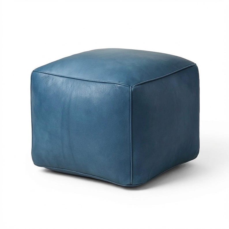 May include: A square, blue leather ottoman. The ottoman is a solid color and has a smooth, slightly textured surface. The edges are neatly stitched, and the overall design is simple and modern. It is a versatile piece of furniture.