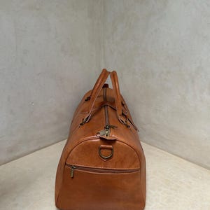 May include: A brown leather duffel bag with a zipper closure and two top handles. The bag has a front zipper pocket and a metal D-ring. The leather has a slightly distressed appearance.