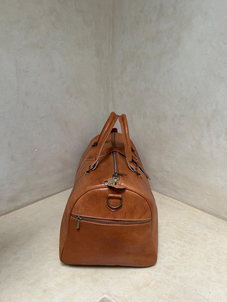 May include: A brown leather duffel bag with a zipper closure and two top handles. The bag has a front zipper pocket and a metal D-ring. The leather has a slightly distressed appearance.