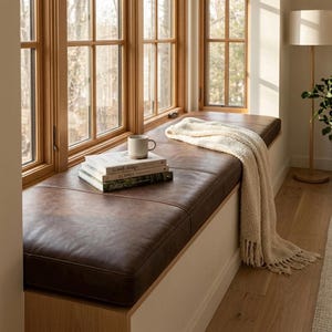 K&ouml;nnte beinhalten: Ein braunes Lederfensterbankkissen mit B&uuml;chern und einer Tasse. Eine cremefarbene Decke ist &uuml;ber dem Kissen drapiert. Der Fensterrahmen ist aus Holz. Die Fensterbank ist 150 cm lang.