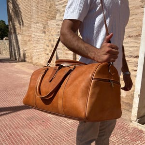 May include: A tan leather duffel bag with a shoulder strap and top handles. The bag has a zipper closure and is carried by a person. The bag is a medium size and appears to be made of high-quality leather.