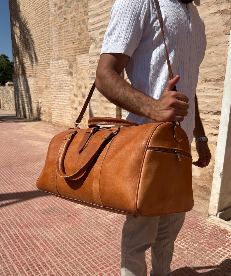 May include: A tan leather duffel bag with a shoulder strap and top handles. The bag has a zipper closure and is carried by a person. The bag is a medium size and appears to be made of high-quality leather.