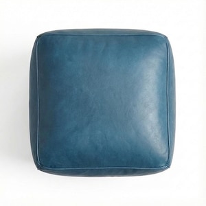 May include: A square, teal-colored leather ottoman. The top and sides are a solid teal color, with a slightly distressed appearance. The edges are neatly stitched, and the ottoman is set against a white background.