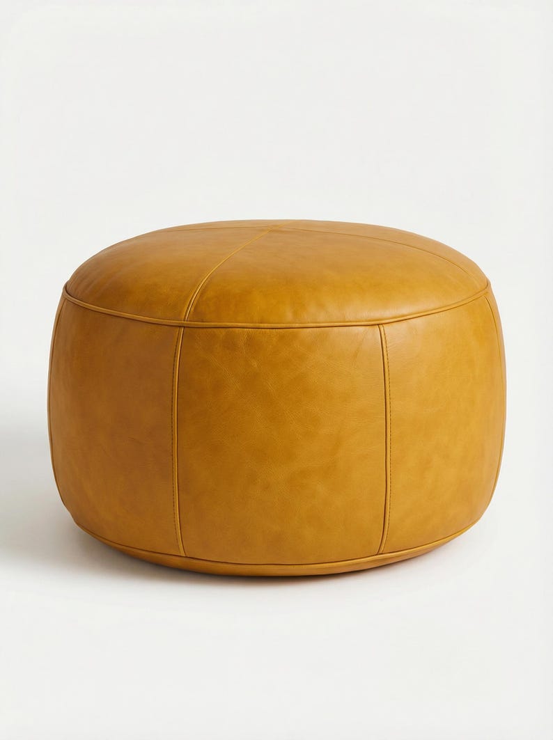 May include: A round, mustard-colored ottoman with a stitched design. The leather-like material has a smooth texture and a warm, inviting hue. The ottoman is a versatile piece of furniture, suitable for various interior styles.