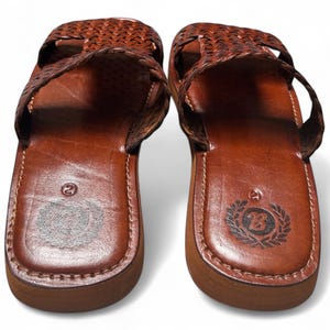 May include: Pair of brown leather sandals with woven straps. The sandals have a rounded toe and a thick sole. The footbed is stamped with a number and a logo. The sandals are a size 9.
