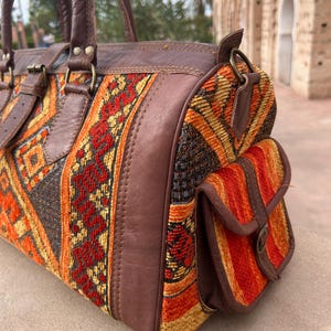 May include: A brown leather and woven fabric duffel bag with a geometric pattern in orange, red, and yellow. The bag has a top handle, side pocket, and leather accents. The bag is approximately 12 inches long.