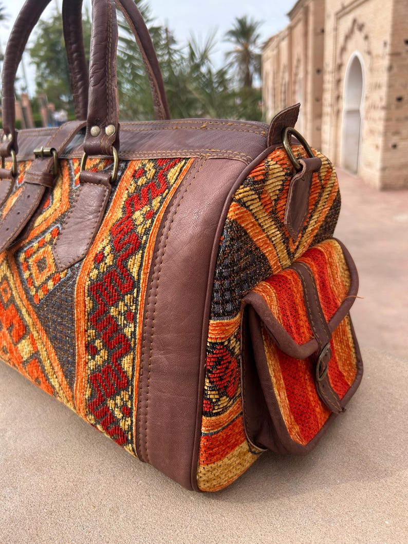 May include: A brown leather and woven fabric duffel bag with a geometric pattern in orange, red, and yellow. The bag has a top handle, side pocket, and leather accents. The bag is approximately 12 inches long.