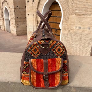 May include: A brown leather and woven fabric travel bag with a top handle and a front pocket. The bag features a colorful geometric pattern in orange, yellow, and black. The bag is sitting on a stone ledge.