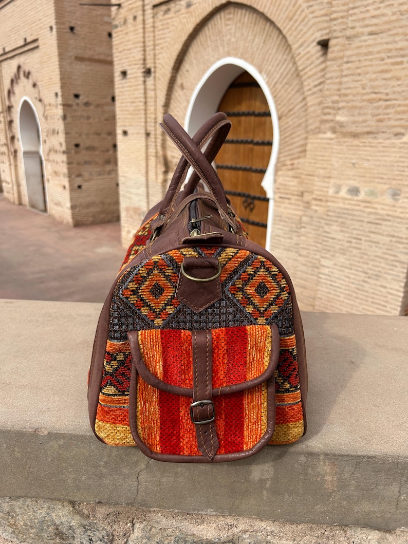 May include: A brown leather and woven fabric travel bag with a top handle and a front pocket. The bag features a colorful geometric pattern in orange, yellow, and black. The bag is sitting on a stone ledge.