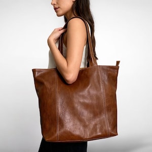 May include: A large, brown leather tote bag with two handles. The bag has a rectangular shape with a flat bottom and a vertical seam down the center. The leather has a slightly distressed appearance.