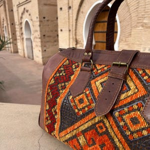 May include: A brown leather and patterned fabric duffel bag with a top handle and shoulder strap. The bag features a geometric design in orange, red, and yellow on a dark background. The bag has a buckle closure and is set against a blurred architectural background.
