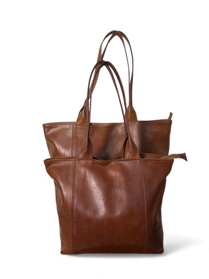 May include: Two brown leather tote bags with long handles. The bags are a medium brown color and appear to be made of a soft, supple leather. The bags have a simple, classic design, with a rectangular shape and a zipper closure.
