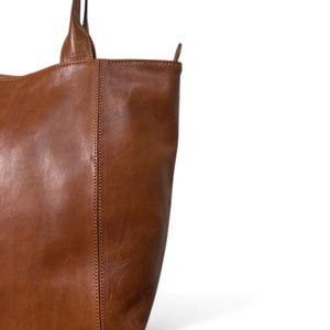 May include: A close-up of a brown leather tote bag. The bag has two handles and a vertical seam down the front. The leather has a natural, textured appearance. The bag is a warm, medium brown color.