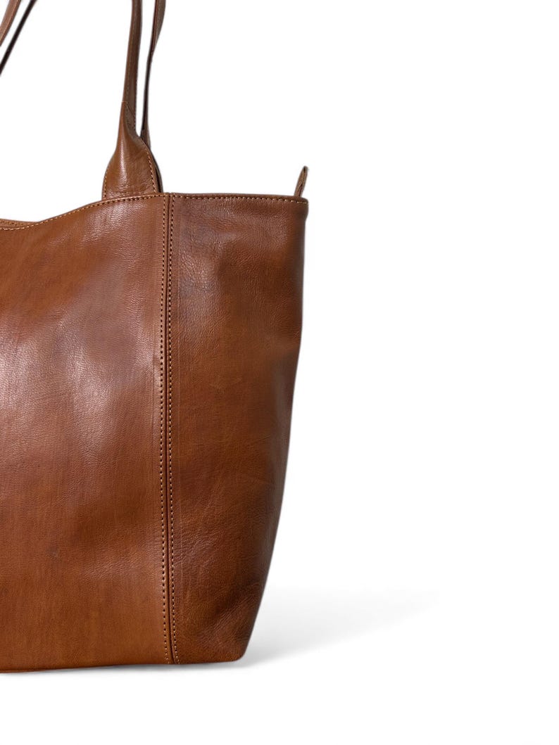 May include: A close-up of a brown leather tote bag. The bag has two handles and a vertical seam down the front. The leather has a natural, textured appearance. The bag is a warm, medium brown color.