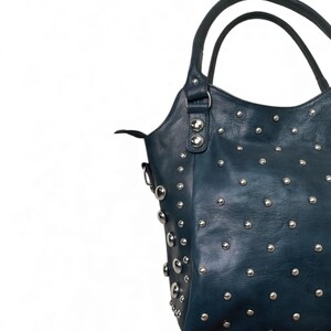 May include: A dark teal handbag with silver-colored studs. The bag has two handles and a shoulder strap. The studs are evenly spaced across the surface of the bag. The bag is made of a textured material.
