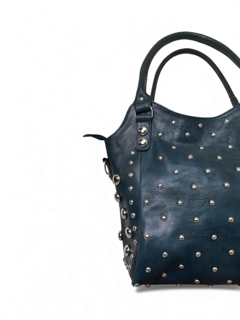 May include: A dark teal handbag with silver-colored studs. The bag has two handles and a shoulder strap. The studs are evenly spaced across the surface of the bag. The bag is made of a textured material.