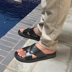 May include: Black leather sandals with crisscross straps and a dark sole. The sandals are worn with light gray pants. The sandals appear to be made of leather and are designed for casual wear.