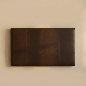 May include: A rectangular, dark brown headboard with a grid pattern of stitched squares. The headboard is mounted on a light beige wall, providing a neutral backdrop. The headboard is made of a smooth, leather-like material.