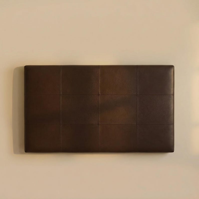 May include: A rectangular, dark brown headboard with a grid pattern of stitched squares. The headboard is mounted on a light beige wall, providing a neutral backdrop. The headboard is made of a smooth, leather-like material.