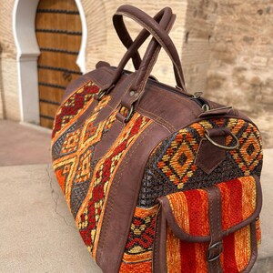 May include: A brown leather and woven fabric duffel bag with a geometric pattern in shades of orange, red, and yellow. The bag has a side pocket, brown leather handles, and a zipper closure. The bag is designed for travel.