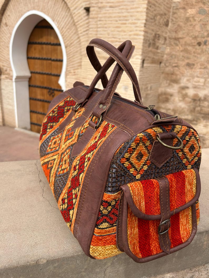 May include: A brown leather and woven fabric duffel bag with a geometric pattern in shades of orange, red, and yellow. The bag has a side pocket, brown leather handles, and a zipper closure. The bag is designed for travel.