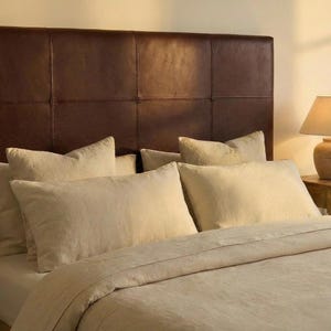 May include: A bed with a brown leather headboard and several beige pillows. The bed is made with a beige comforter and sheets. A lamp is visible on the right side of the bed.