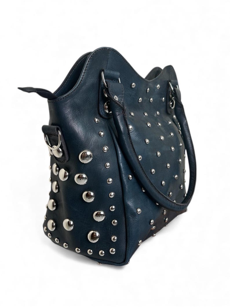 May include: A teal handbag with silver-colored studs. The bag has a structured shape with a top handle and a shoulder strap. The studs are evenly spaced across the front, sides, and back of the bag, adding a decorative element to the design.